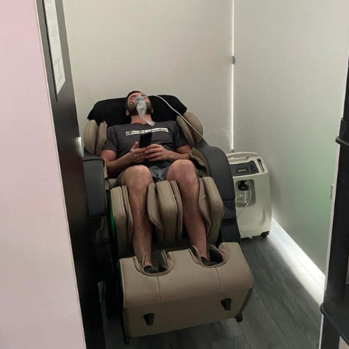 A man reclining in a beige massage chair with his legs extended, wearing a face mask and holding a phone in a spa-like room with a small machine beside the chair.